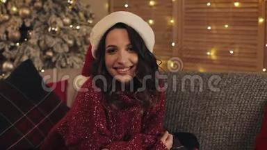 放大年轻美丽的黑发女子，戴着桑塔帽，坐在圣诞树附近的沙发上，闪闪发光的灯光
